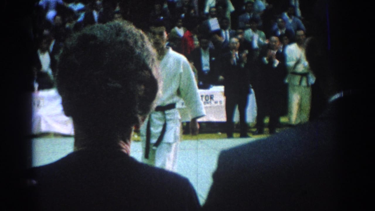 Foreground silhouettes face competitors in white uniforms on a mat before a seated audience.