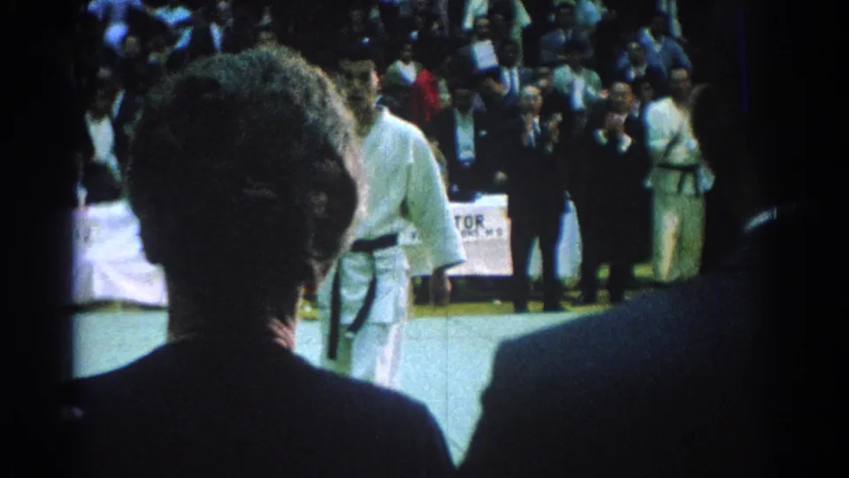 1966 San Gabriel frame with spectators seen from behind while martial arts competitors in white gi stand on the mat.