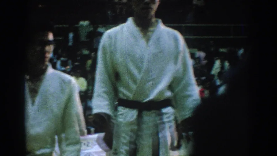In San Gabriel, California, 1966, karate competitors in white gi uniforms stand near a crowded indoor ceremony area.