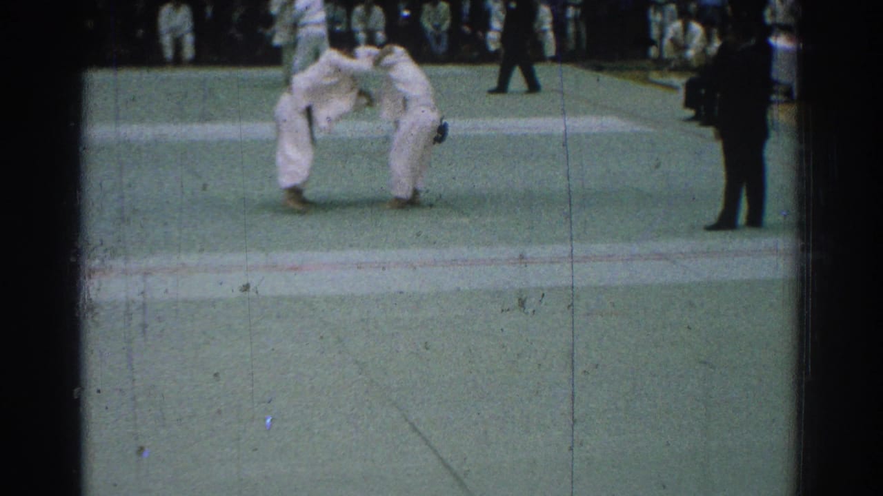 Two judo competitors in white gi grapple on a green mat with spectators behind them.