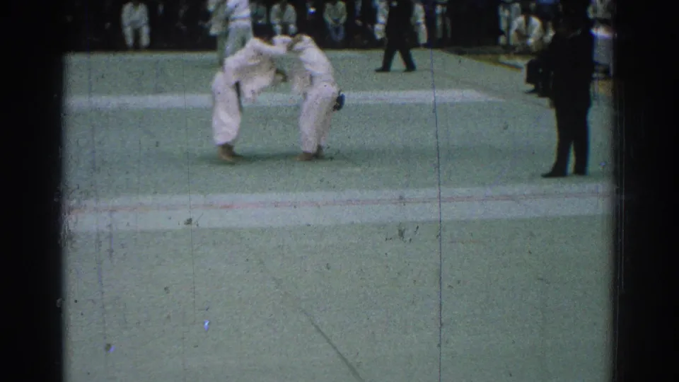 In 1966 San Gabriel, two martial artists in white gi face each other on a green indoor mat as uniformed spectators watch from the back.