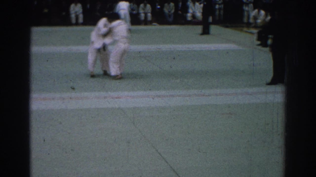 Two judo competitors hold each other at center mat while spectators sit in the background.