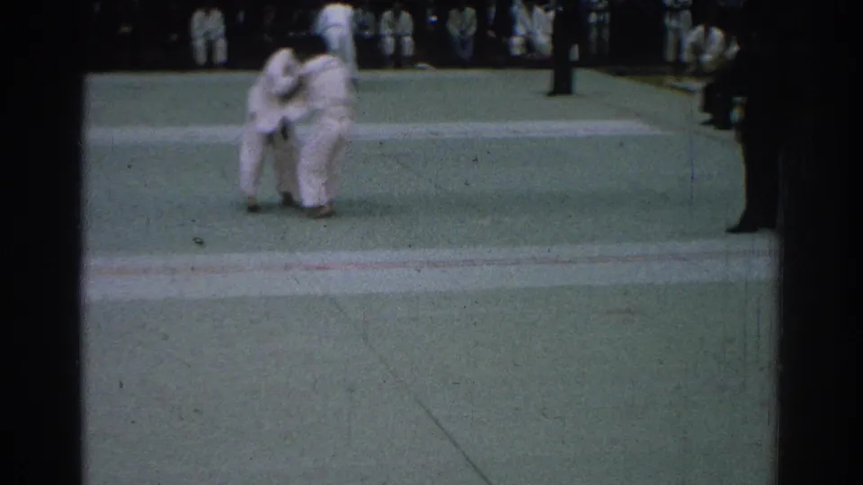 The San Gabriel judo match continues as the two competitors clinch on the mat, framed by the dark film gate and distant spectators.