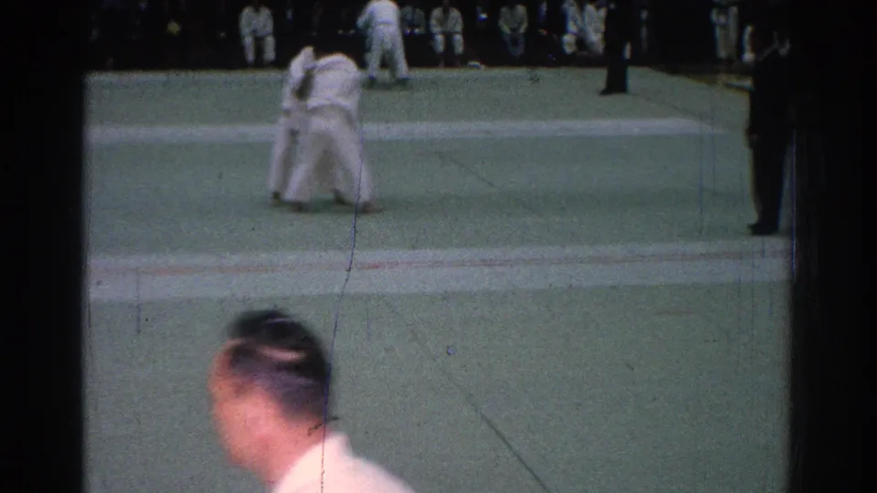During the 1966 indoor judo match, the camera catches the competitors in a close hold while a foreground spectator briefly crosses the frame.