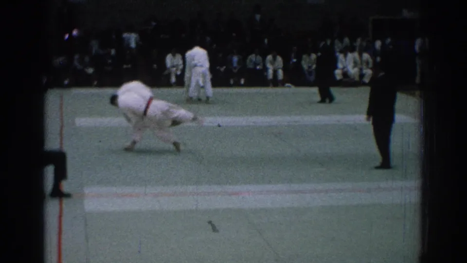 A judo throw or takedown is visible on the green mat, with one competitor low and extended as spectators watch in the San Gabriel gym setting.