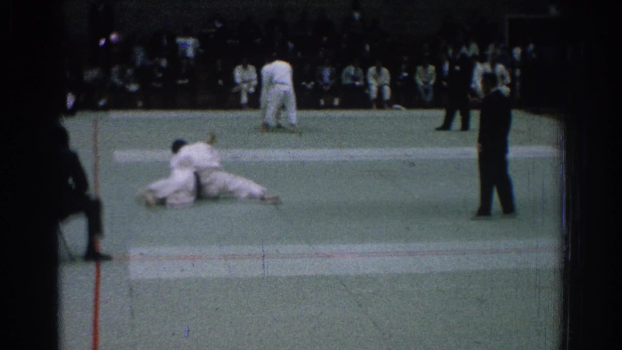 In San Gabriel, California in 1966, two men in white judo uniforms are low on a green mat during a match with a suited referee nearby.