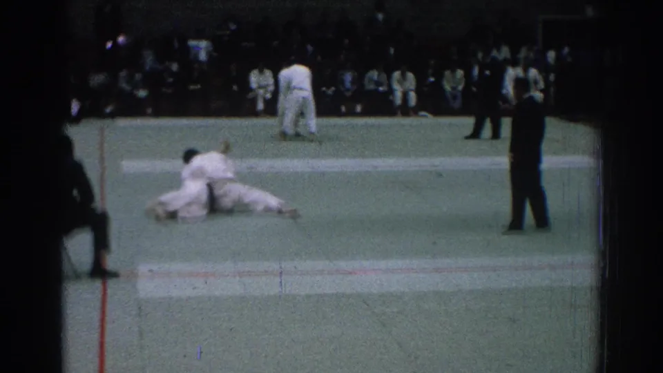 In San Gabriel, California in 1966, two men in white judo uniforms are low on a green mat during a match with a suited referee nearby.
