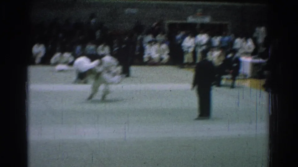 At the end of the 1966 San Gabriel judo clip, two competitors stand and grip on the green mat while a suited referee watches.
