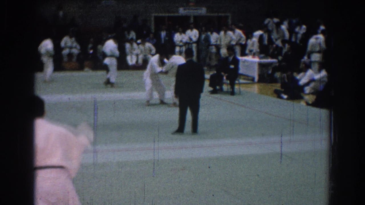 Two martial artists grapple on the mat while a referee observes and spectators sit along the side.