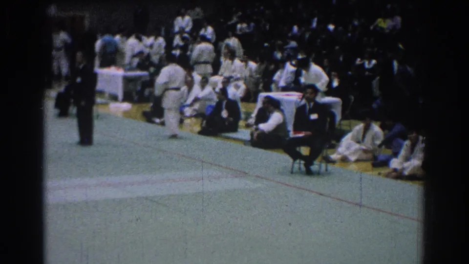 The closing frame shows the side of the martial arts event, with seated participants, spectators, tables, and the edge of the competition area.
