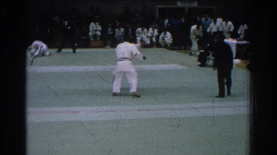 1966 San Gabriel home-movie frame showing two karate competitors on a green mat with a dark-suited referee and seated spectators.