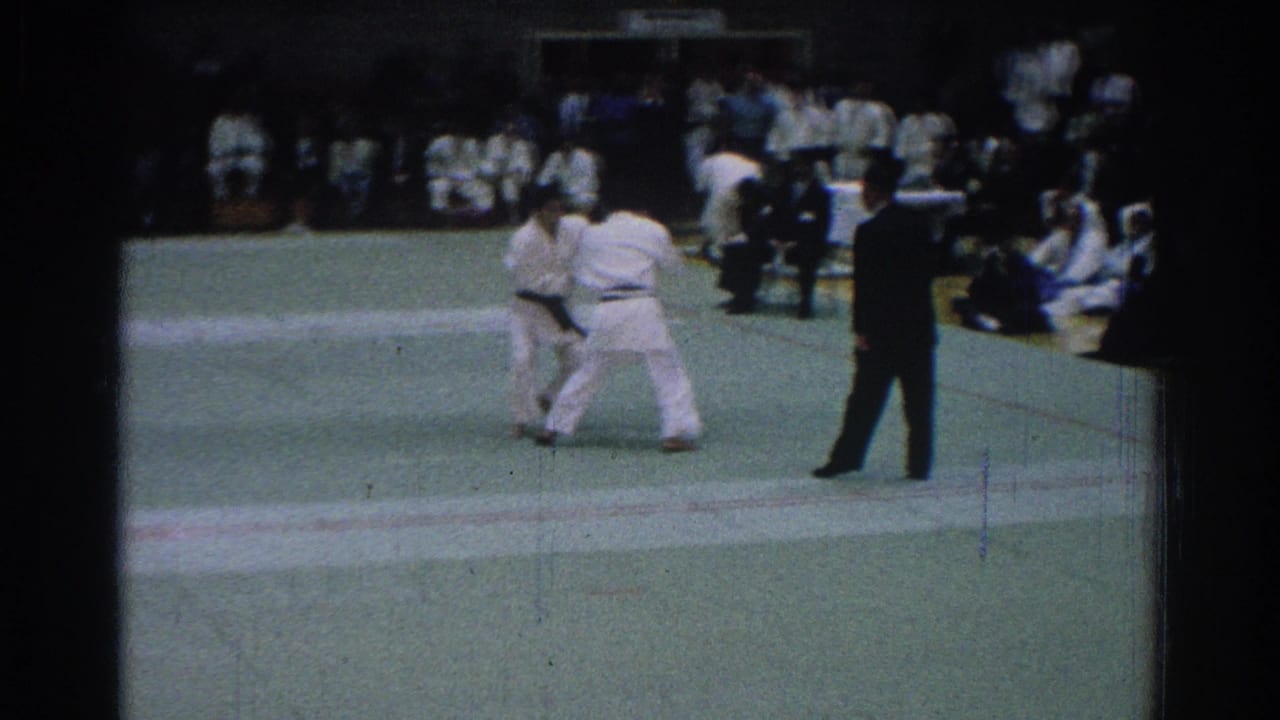 Two karate competitors in white gi grapple on the mat while a referee observes from the right.