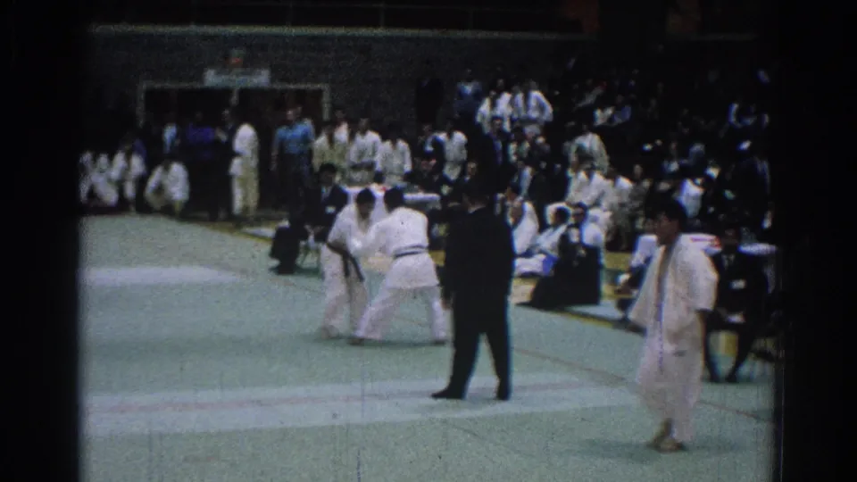 Indoor 1966 San Gabriel karate competition frame with two competitors squared up, a suited referee, and spectators seated around the mat.