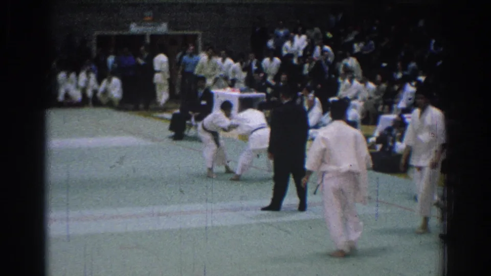 1966 San Gabriel judo competitors in white gi face each other on a green mat as a dark-suited referee stands nearby.