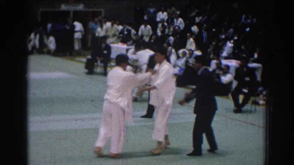 At a San Gabriel judo match in 1966, two athletes in white gi grip each other while the referee moves close to observe.