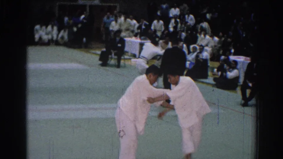 A close 8mm view shows two judo athletes gripping near the foreground during a 1966 San Gabriel competition.