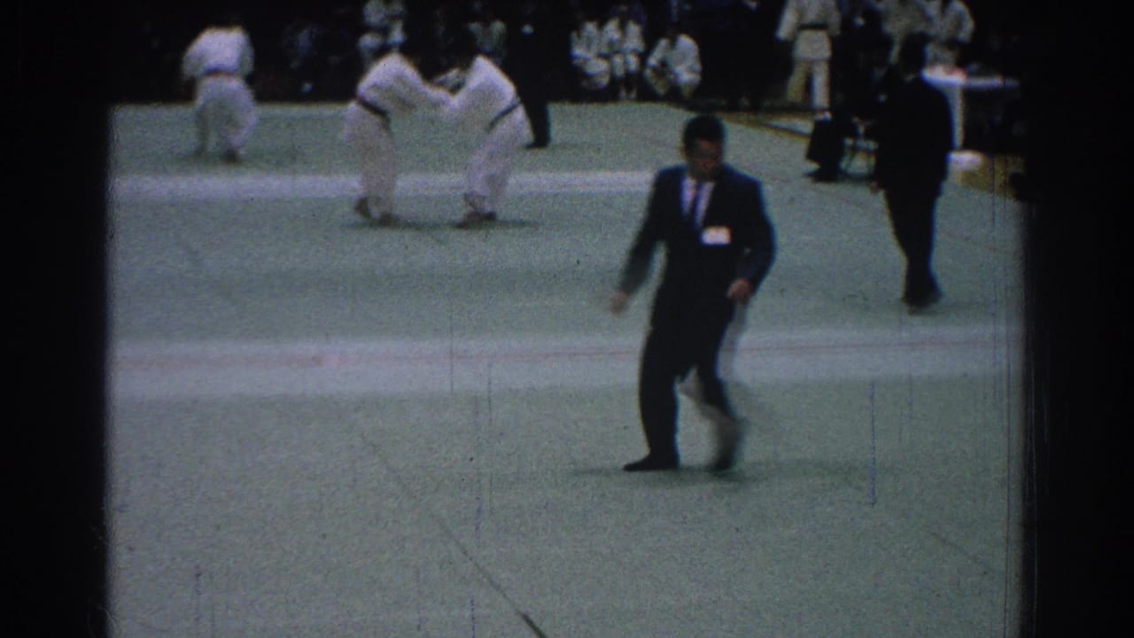 A suited referee walks across the mat as two fighters grapple in the background.