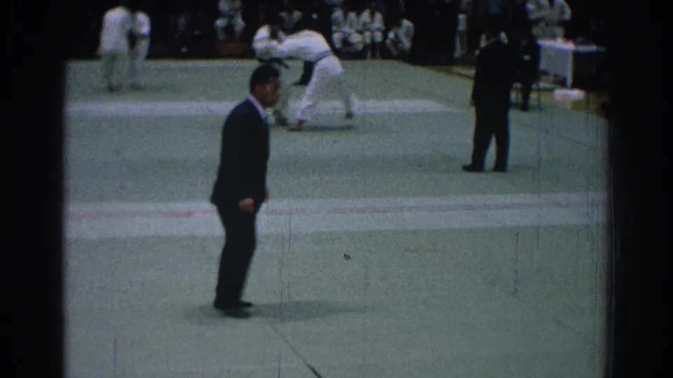 1966 San Gabriel home-movie frame showing a referee near the foreground and jujitsu competitors in white gi grappling in the background.