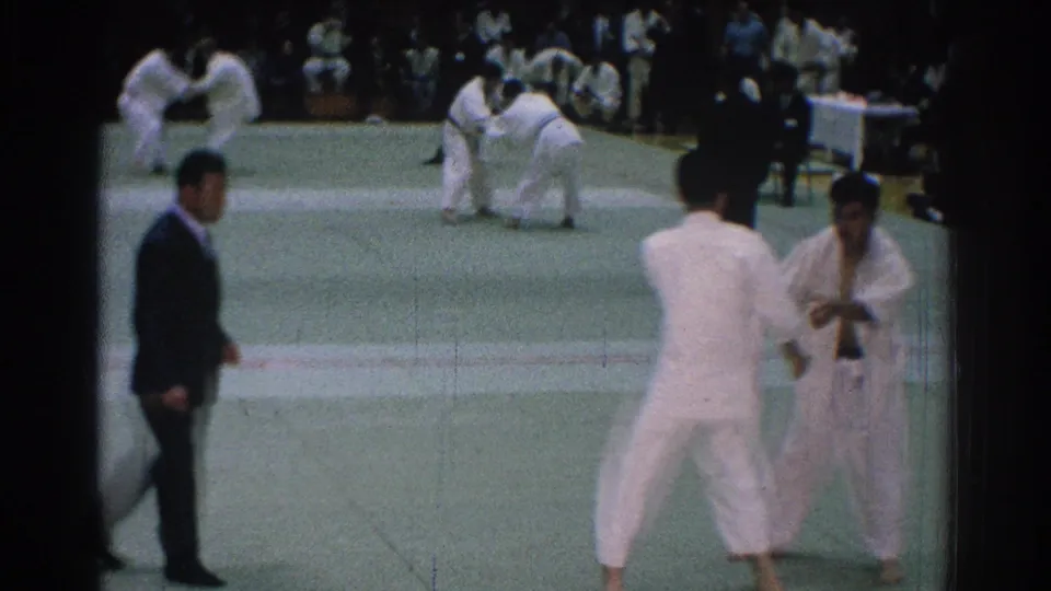 1966 San Gabriel 8mm frame with white-uniformed fighters in the foreground and another grappling match visible across the green mat.