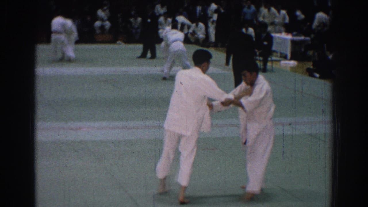 Two judo competitors in white gi face off on a green mat during a 1966 San Gabriel competition, with officials and other matches behind them.