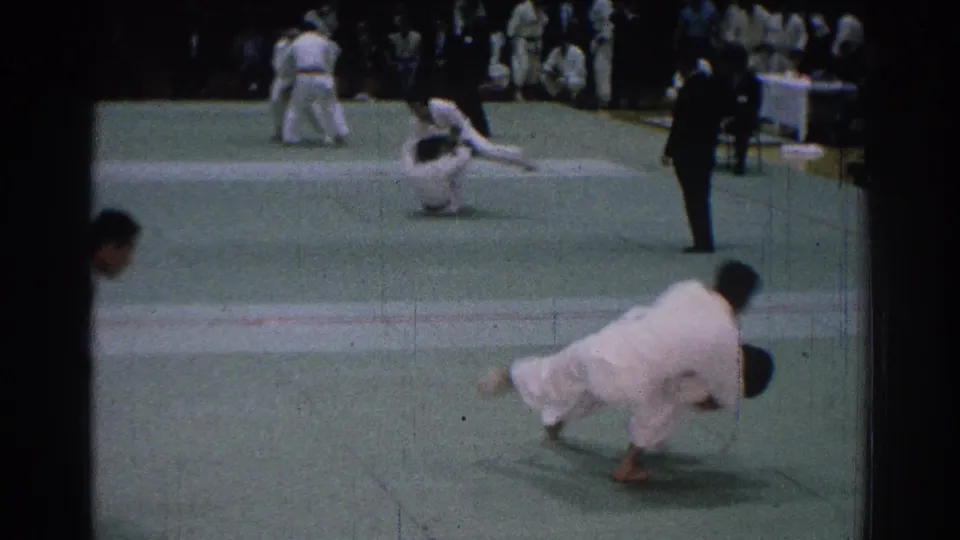 A competitor drives into a throw or takedown during the San Gabriel judo meet, with officials and spectators visible beyond the mat.