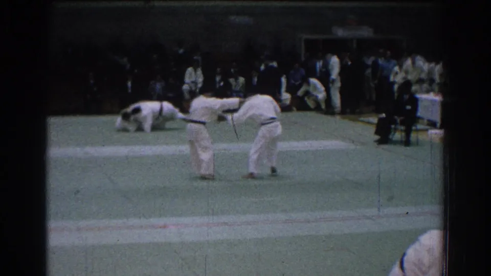 Two judo competitors bow on the green mat during the 1966 San Gabriel event, while other athletes and spectators remain in the background.