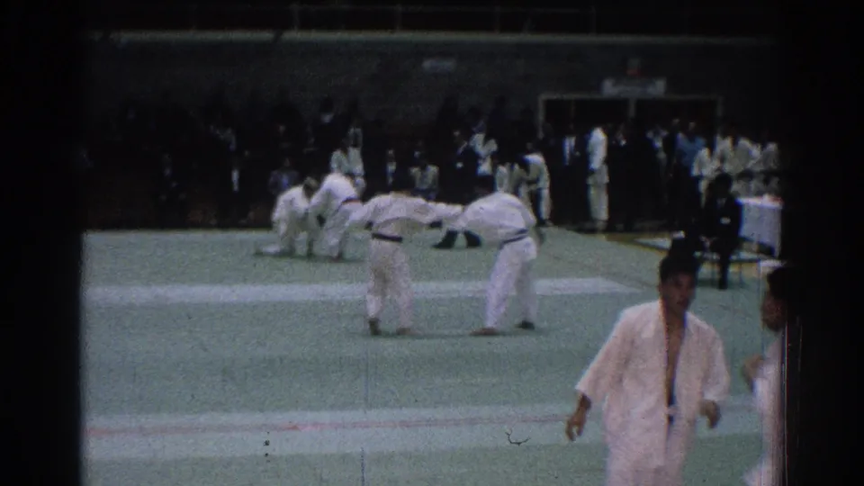 Competitors bow on the mat at the San Gabriel judo competition, with a foreground figure passing close to the camera.