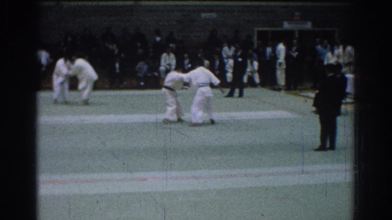 1966 San Gabriel judo match with two men in white gis grappling on a green mat before seated spectators and a dark-suited official.
