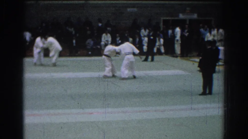 1966 San Gabriel judo match with two men in white gis grappling on a green mat before seated spectators and a dark-suited official.