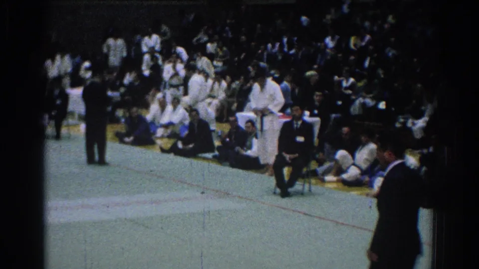 Sideline view of the San Gabriel judo event with seated spectators, competitors in white gis and dark-suited officials around the mat.