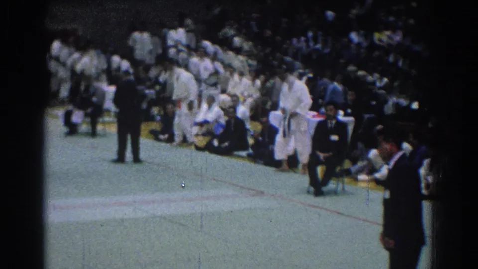 San Gabriel 1966 home-movie frame showing karate competitors in white gi near dark-suited judges and seated spectators around a green mat.