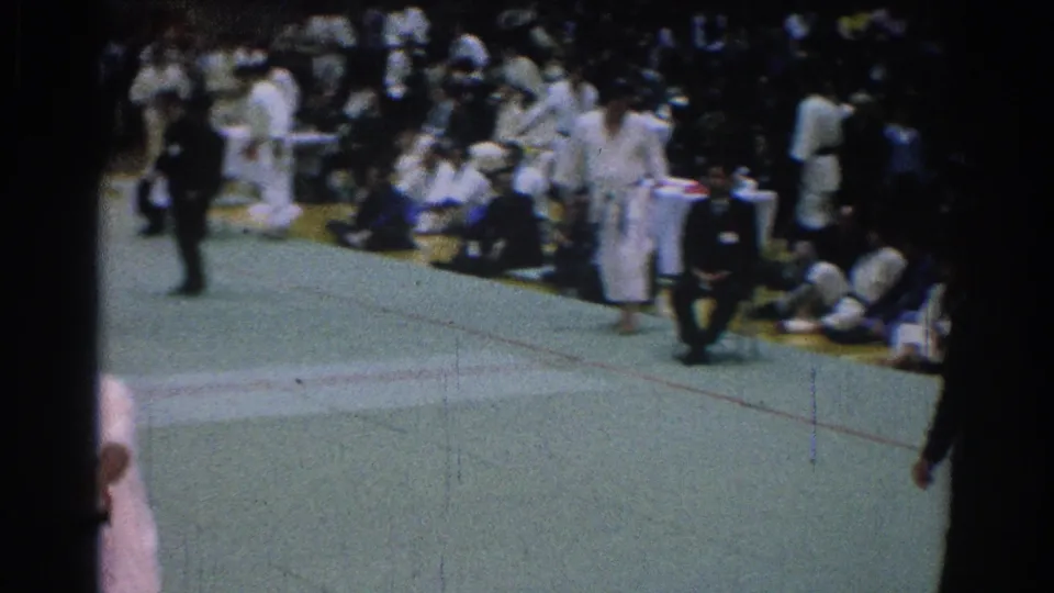 San Gabriel 1966 home-movie frame showing a dark-suited official crossing the green mat as karate athletes and spectators wait nearby.
