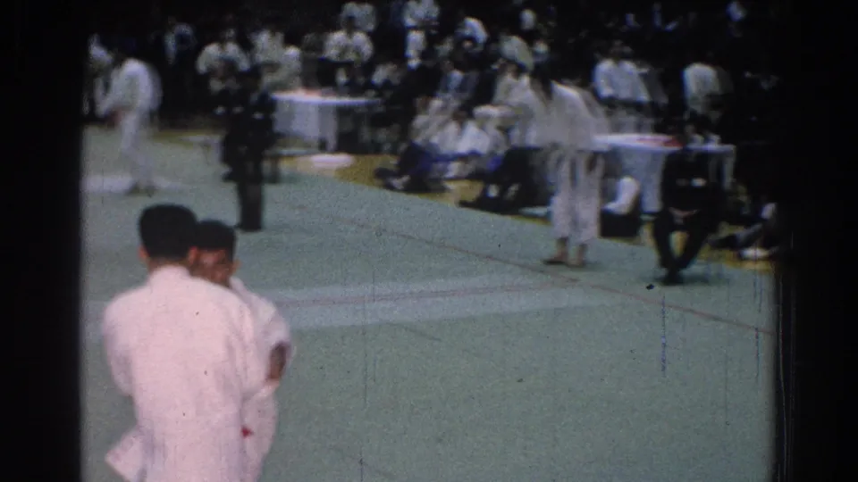 San Gabriel 1966 frame with two white-gi karate competitors in the foreground and judges, spectators, and other athletes around the mat.