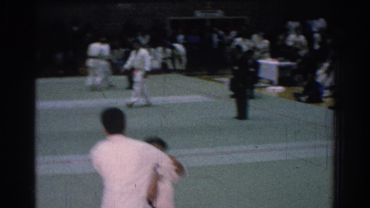 1966 San Gabriel home-movie frame showing two judo competitors in white gi on a green mat with seated spectators behind them.