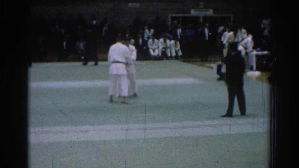 1966 San Gabriel judo match frame with two competitors in white gi on a green mat and spectators seated along the back wall.