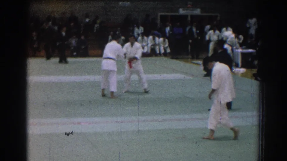 1966 San Gabriel frame showing two judo competitors on the mat with a referee nearby and a white-gi participant crossing in front.