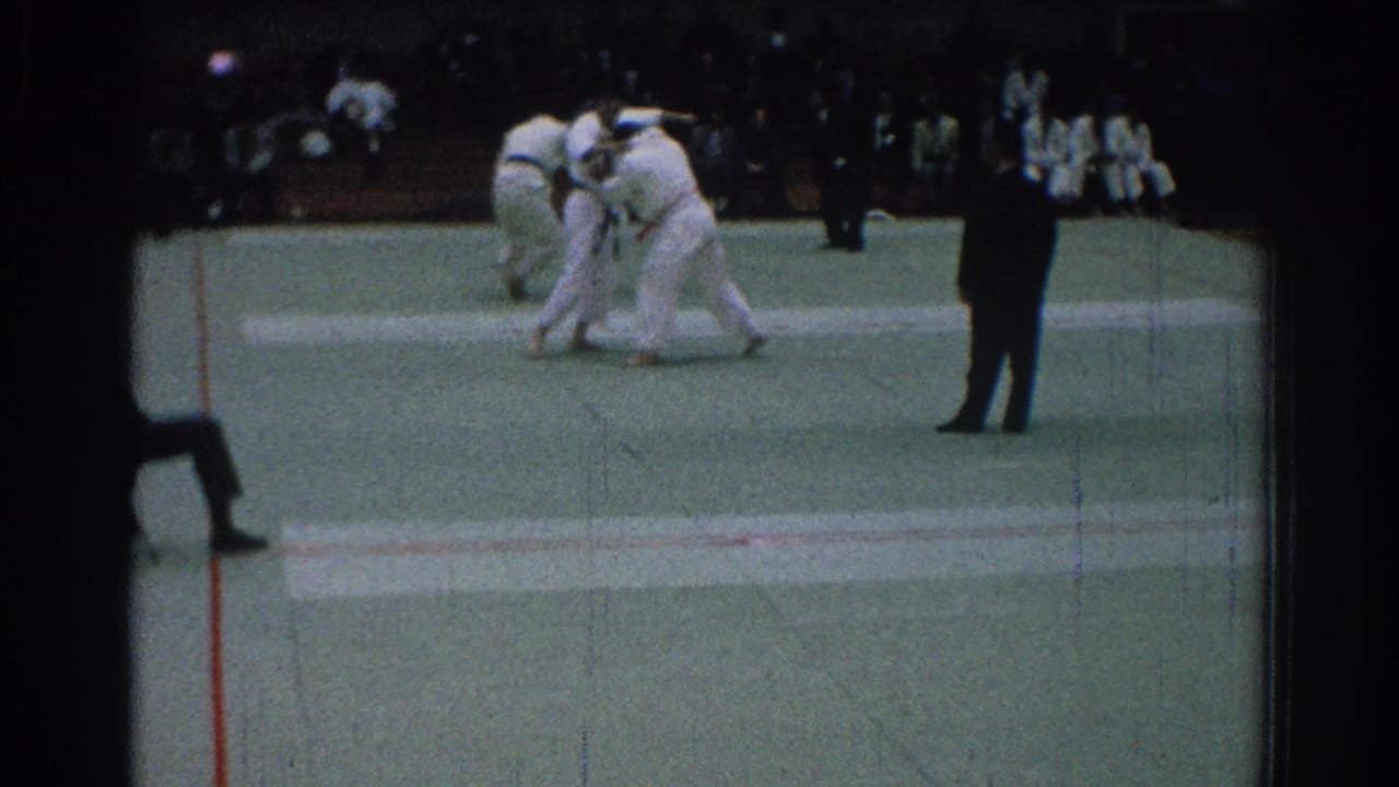 Two karate competitors grapple on a green mat as a referee stands nearby.