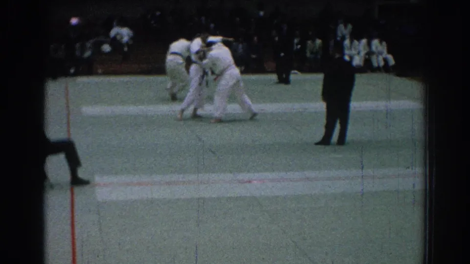 1966 San Gabriel footage frame with two karate competitors in white gi grappling while a dark-suited referee watches.