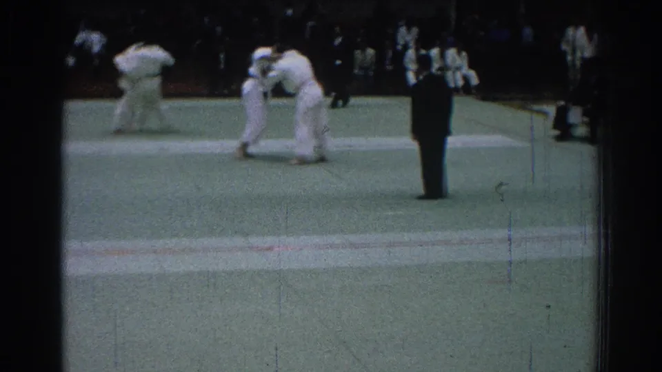 1966 San Gabriel home-movie frame showing karate competitors in white gi engaged on the mat with a referee observing.