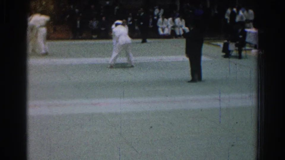 1966 San Gabriel frame showing white-clad karate competitors moving on a green mat as spectators sit in the background.