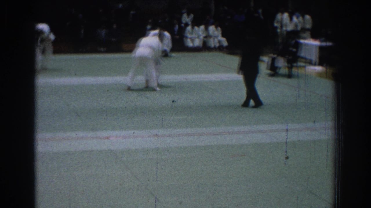 Two judo competitors grapple on a green mat while a referee stands nearby.