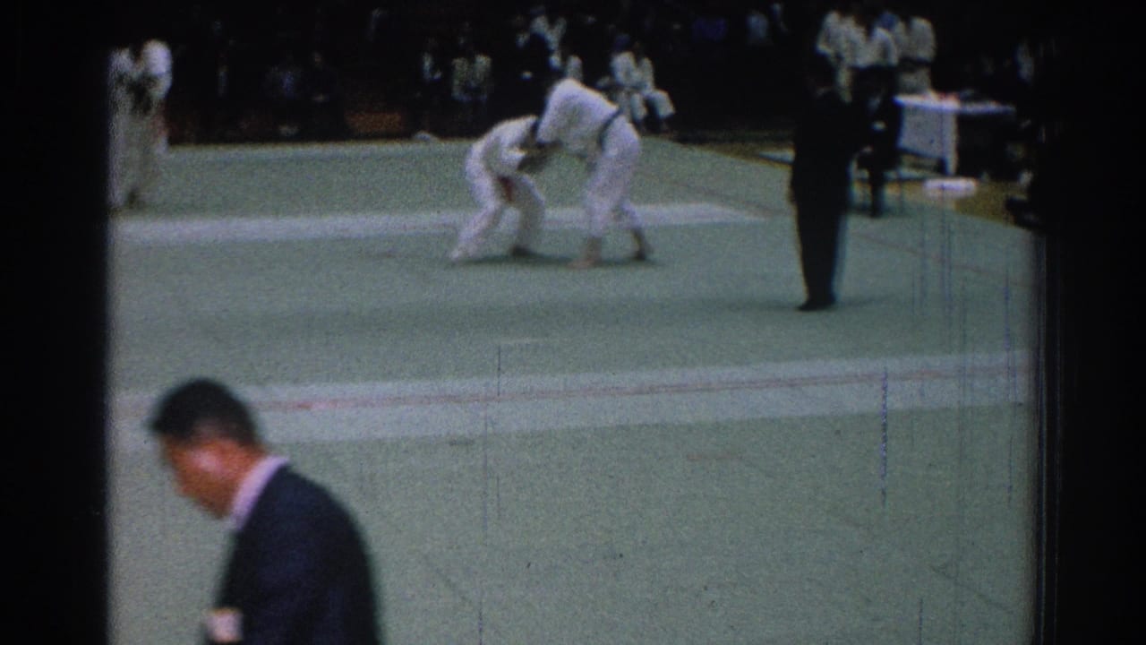 Judo competitors grapple in the distance as a suited man passes in the foreground.