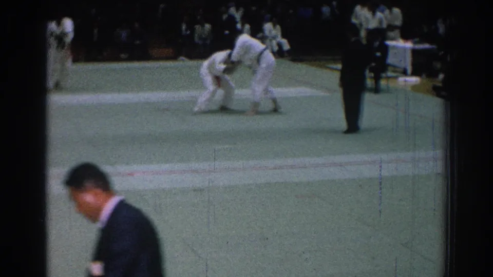 A suited man crosses the foreground while two judo competitors continue grappling on the mat in San Gabriel, California.