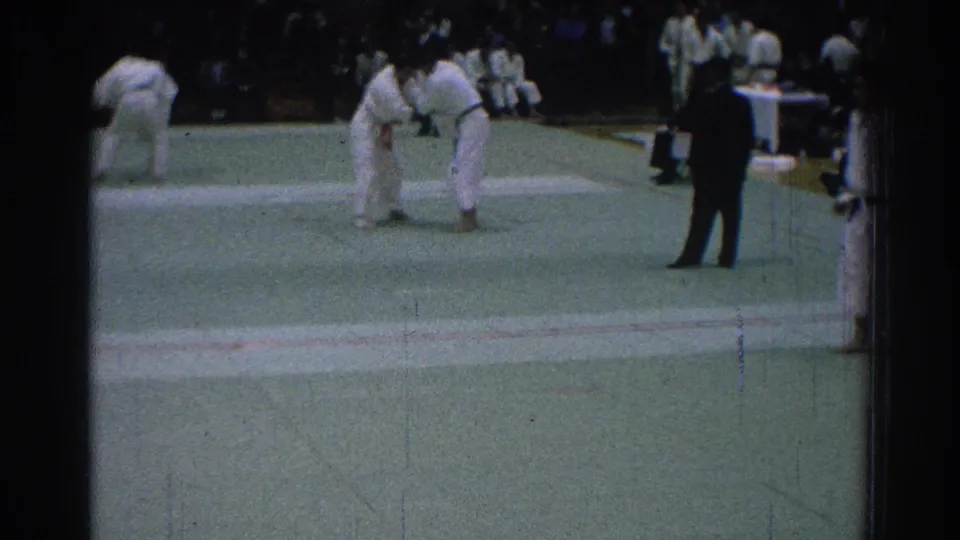 Two judo competitors face off on the green mat during the 1966 San Gabriel tournament as a referee stands to the side.