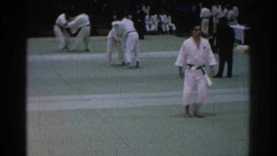 A competitor in a white gi walks near the camera as another judo exchange continues behind him on the mat.
