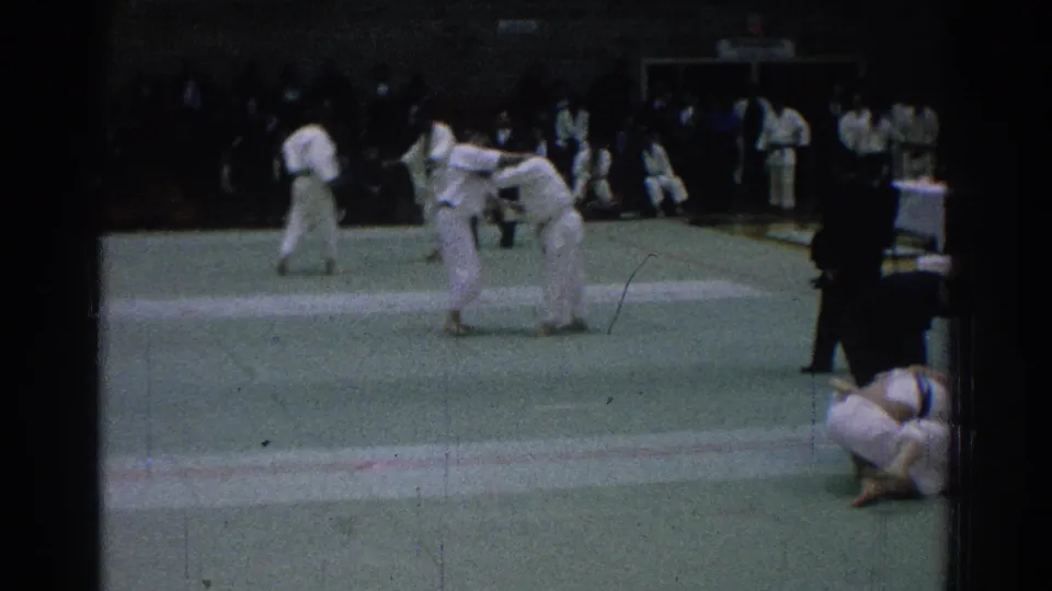 In 1966 San Gabriel, two white-uniformed martial arts competitors engage on a green mat while spectators and other participants watch from the sideline.