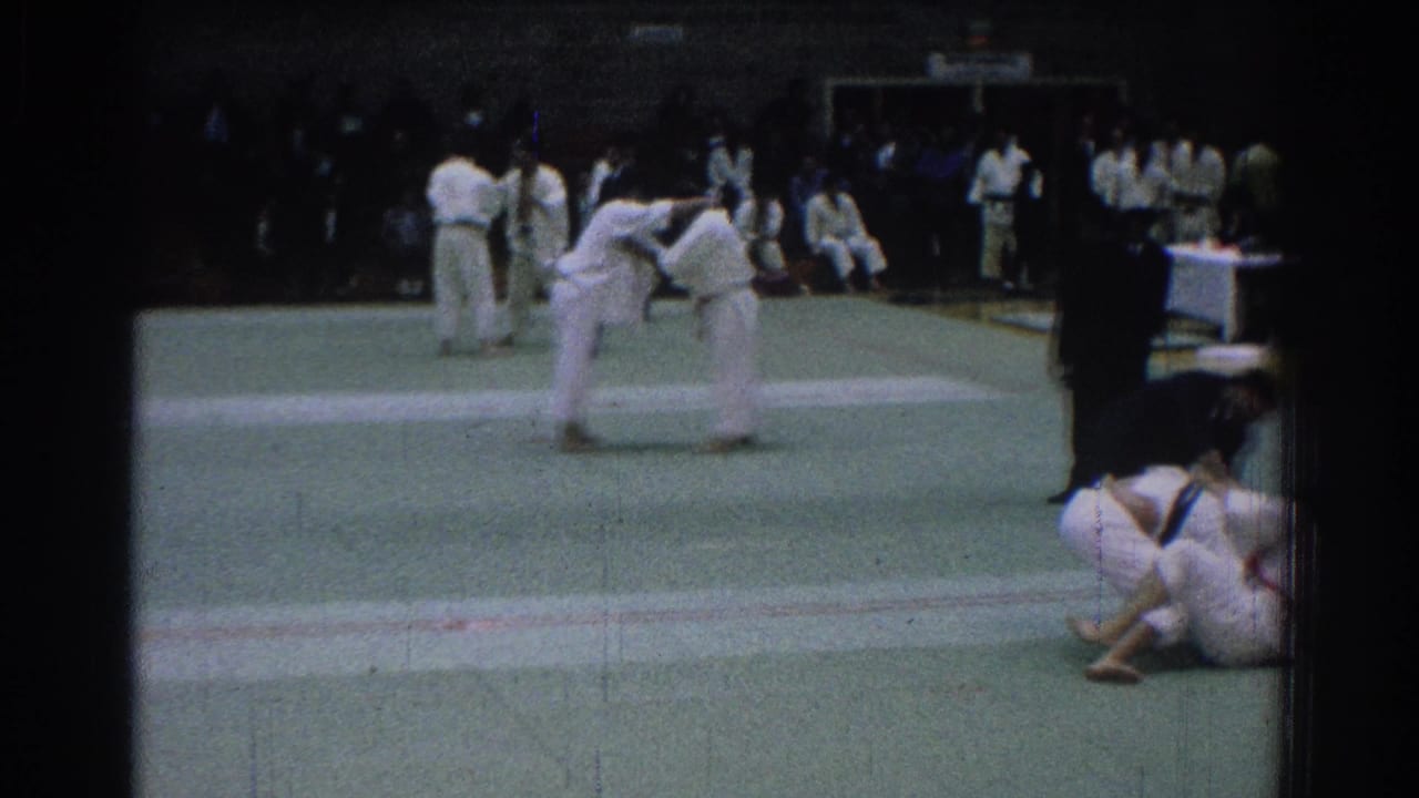 A martial arts pair grapple near center mat as other uniformed participants stand nearby.