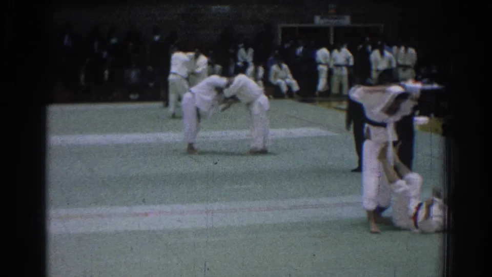 The 1966 gym footage shows a close grappling exchange on the mat, with additional white-uniformed participants and observers gathered around the competition area.