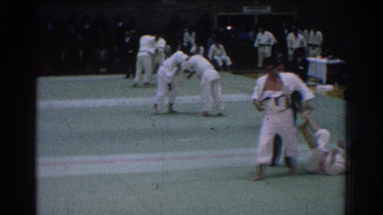 Competitors in white gi uniforms grapple on a green mat as others wait nearby.