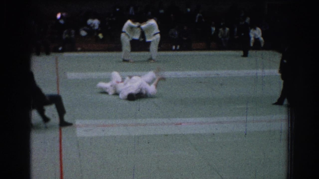 Two martial arts competitors lie on the mat during a bout with judges nearby.